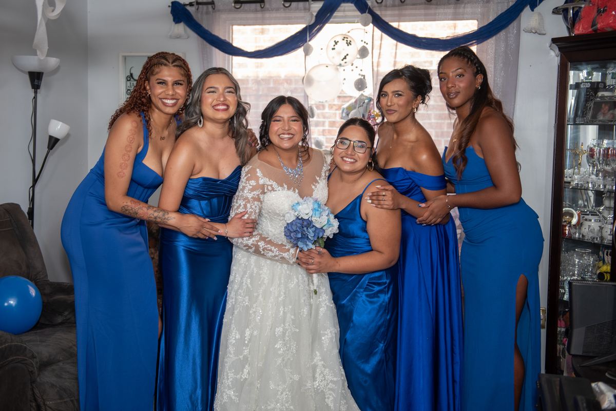 A bride in a white lace gown and her bridesmaids in royal blue gowns stand together in a decorated space with blue drapery.