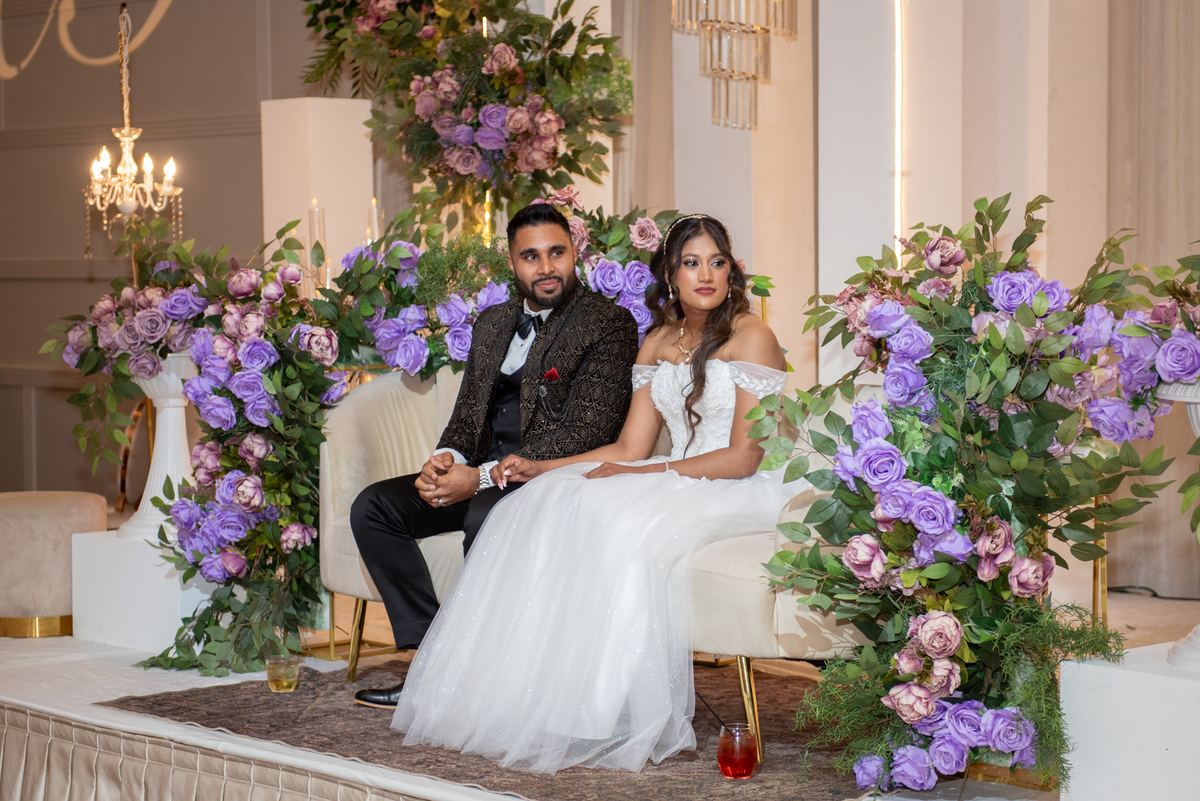 A newlywed couple in formal attire sit on an ornate bench, surrounded by floral arrangements and chandeliers, holding hands.