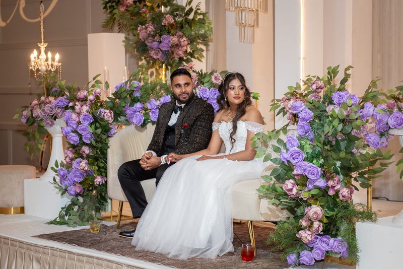A newlywed couple in formal attire sit on an ornate bench, surrounded by floral arrangements and chandeliers, holding hands.