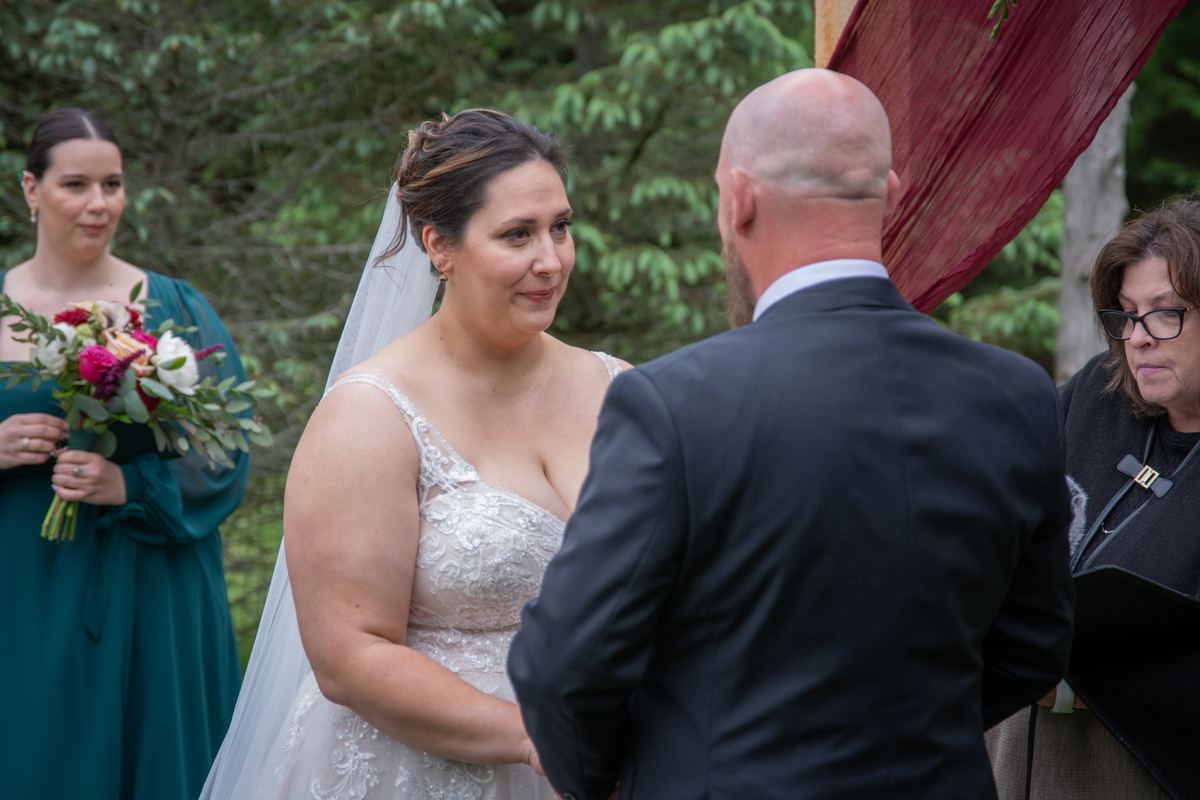 Bride in lace gown smiles at groom during outdoor ceremony under red canopy, with bridesmaid and officiant present.