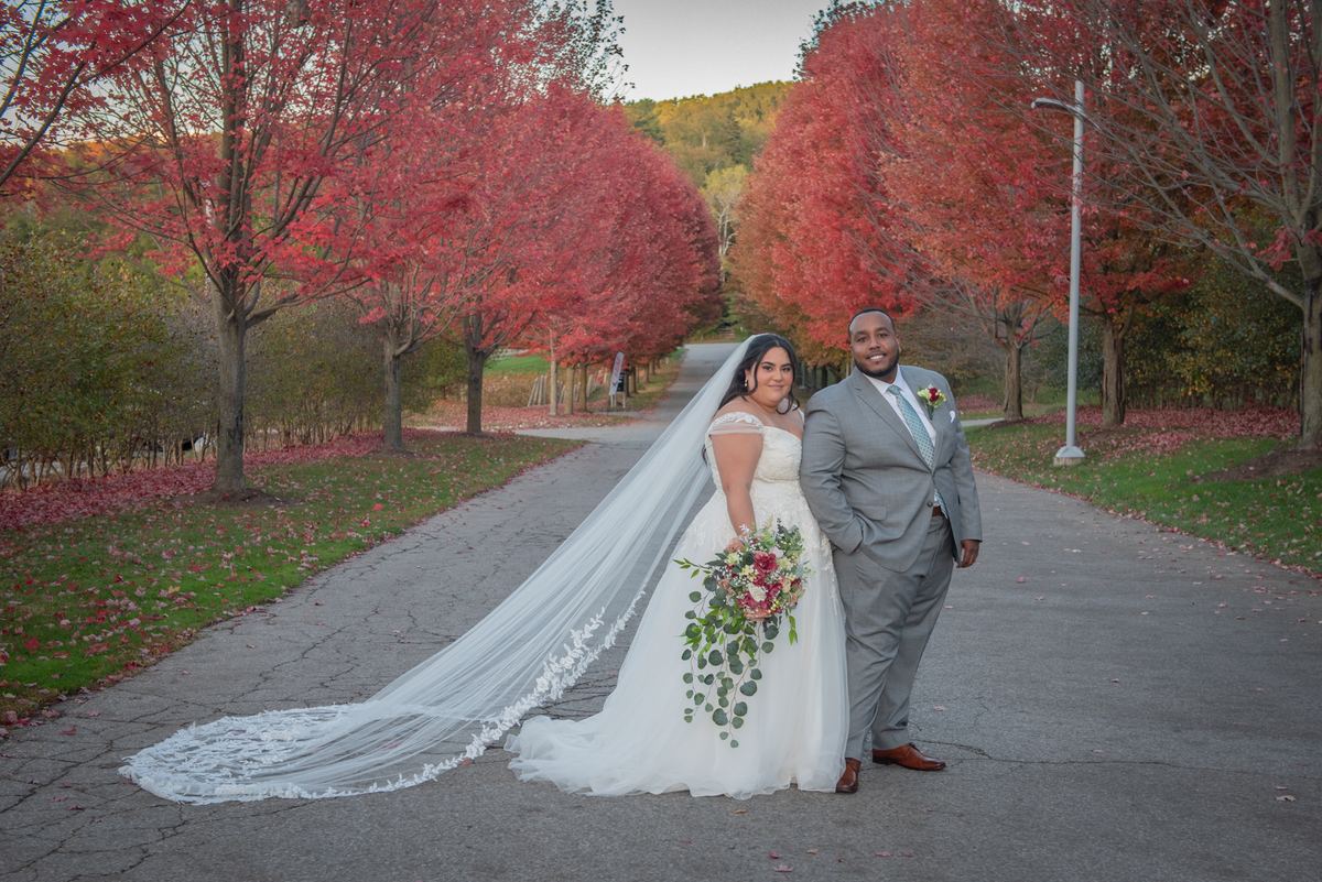 A smiling bride in a white dress and floral bouquet stands with her groom in a gray suit among autumn trees.