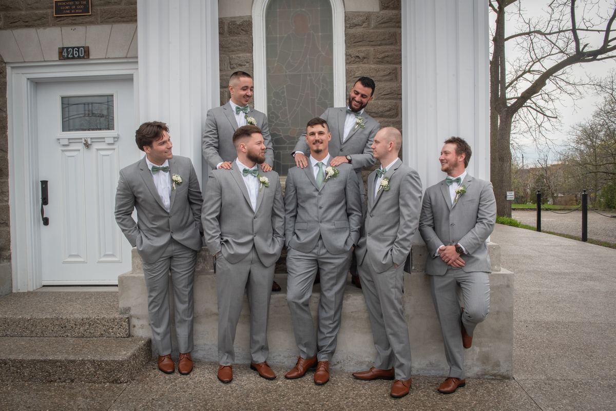 Groomsmen in matching gray suits and green bow ties laugh together on stone steps outside a church with white columns.