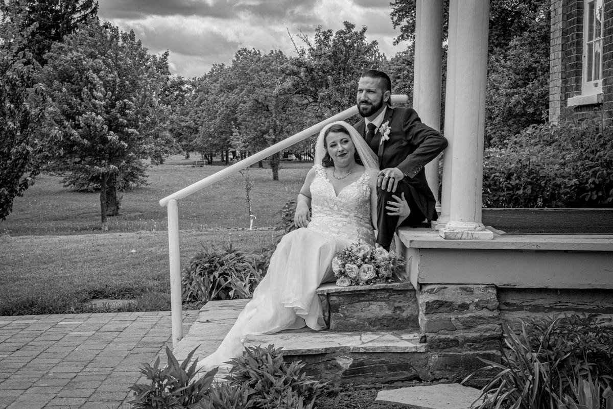 Bride and groom sit happily on stone steps with trees and garden foliage surrounding them, both looking toward the camera.