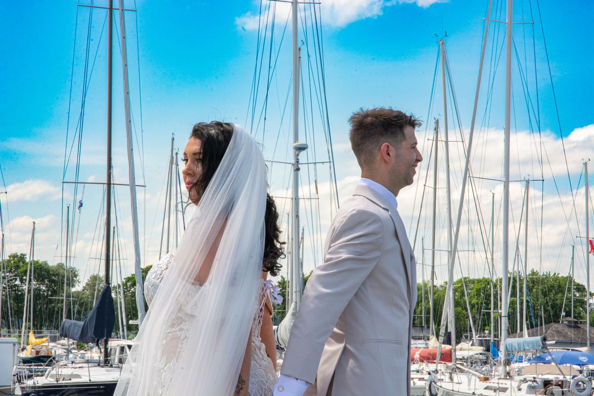 A bride and groom stand beside sailboat masts under a bright blue sky during a marina wedding photoshoot.