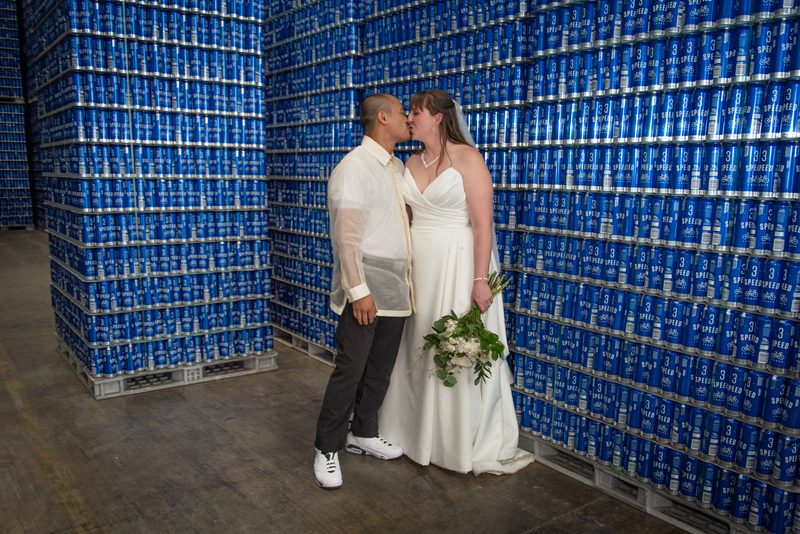 A bride and groom kiss in an industrial warehouse filled with blue beer cans, illuminated by soft lighting.