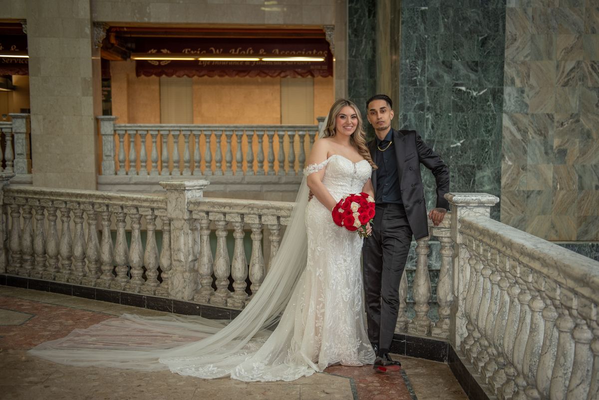 A bride in a white lace gown and veil stands with her groom on an ornate marble balcony, holding red roses.