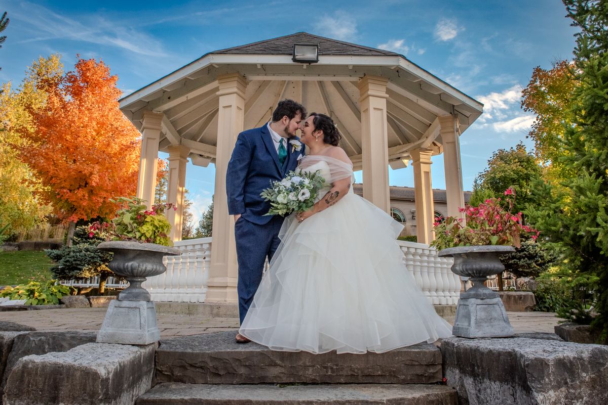 A bride and groom kiss on stone steps under a blue sky, surrounded by autumn foliage and decorative urns.