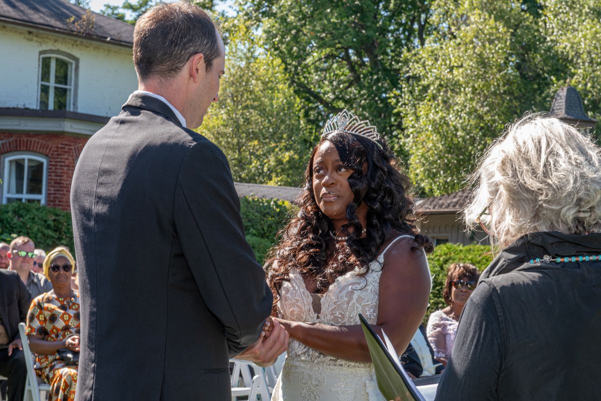 A bride in a white lace gown and tiara exchanges vows with her groom, hands clasped, under dappled sunlight from lush trees.