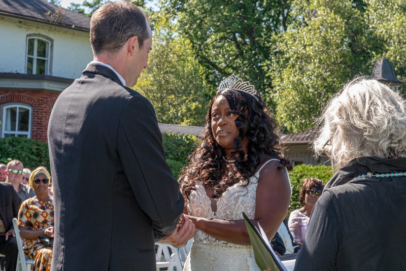 A bride in a white lace gown and tiara exchanges vows with her groom, hands clasped, under dappled sunlight from lush trees.