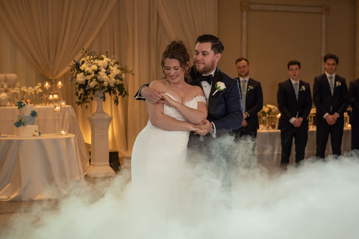 A bride and groom sway in their first dance wrapped in swirling white fog, candles and formally dressed guests behind them.