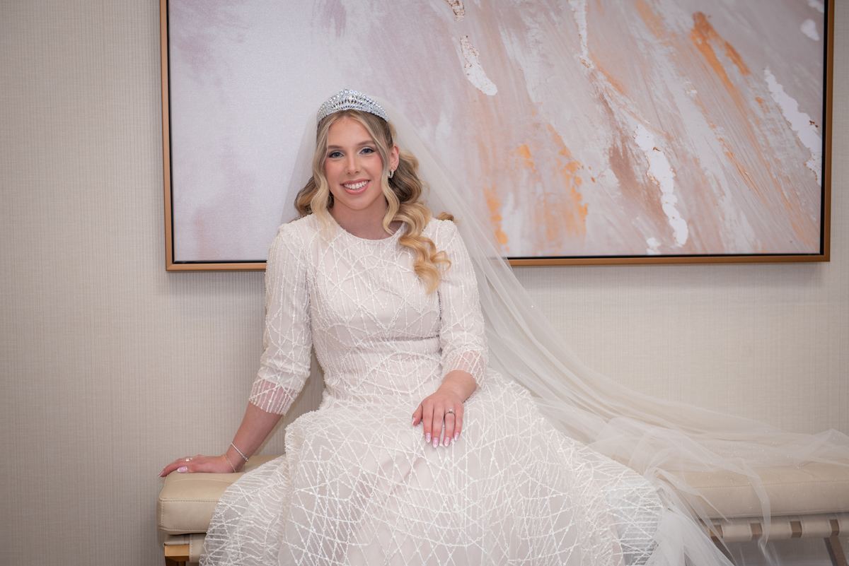 A smiling bride in a white gown and veil sits on a bench near an abstract painting, her dress catching the ambient light.