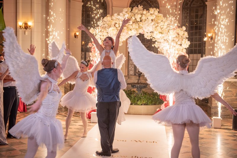 A bride in angel wings dances down an aisle flanked by bridesmaids as fireworks illuminate the hall.