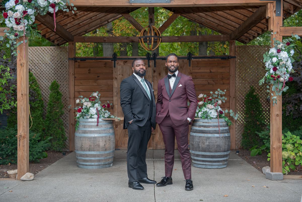 Two men stand under a floral-adorned wooden arbor flanked by decorated wine barrels in soft daylight.