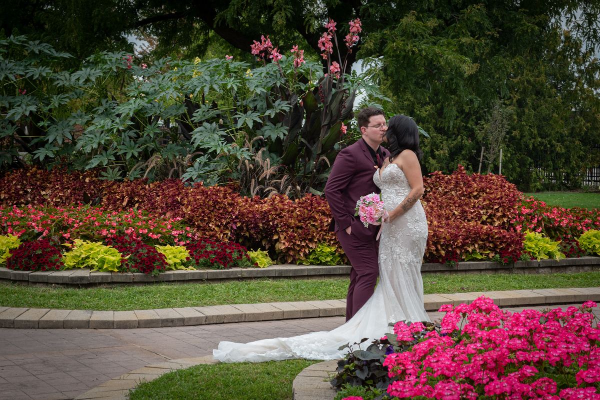 A bride in white and groom in maroon suit kiss in a garden filled with pink and red flowers, bathed in soft daylight.