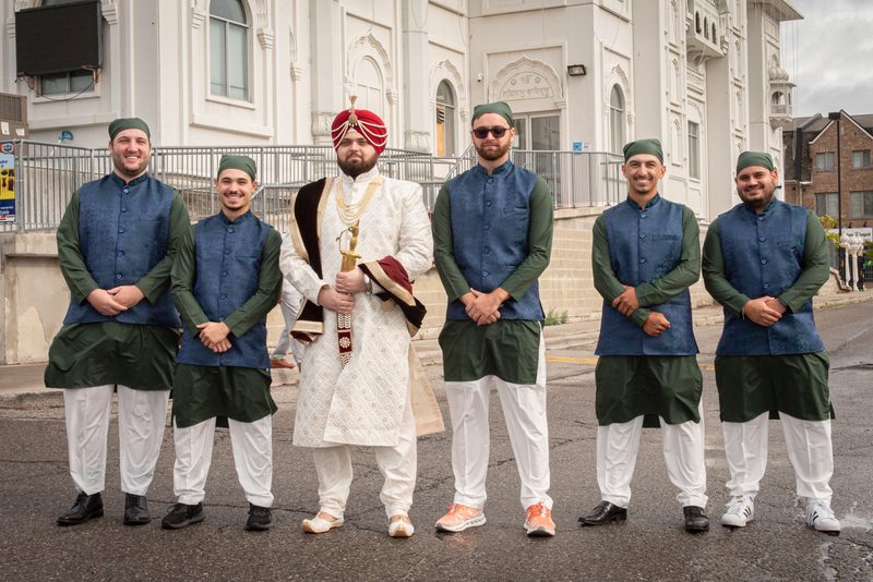 A Sikh groom in white with a red turban and sword stands with his groomsmen in front of a white Gurdwara.