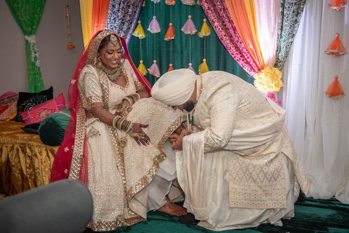 Bride in cream and gold lehenga with red dupatta smiles as groom in sherwani touches her feet, surrounded by festive decor.