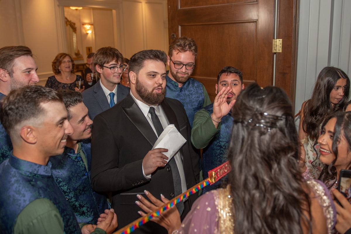 A groom in a black tuxedo speaks to smiling guests at a festive event with colorful decorations and formal attire.