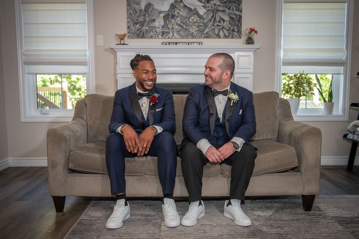 Two grooms in navy tuxedos laugh together on a beige sofa in a cozy room with a fireplace and family sign.