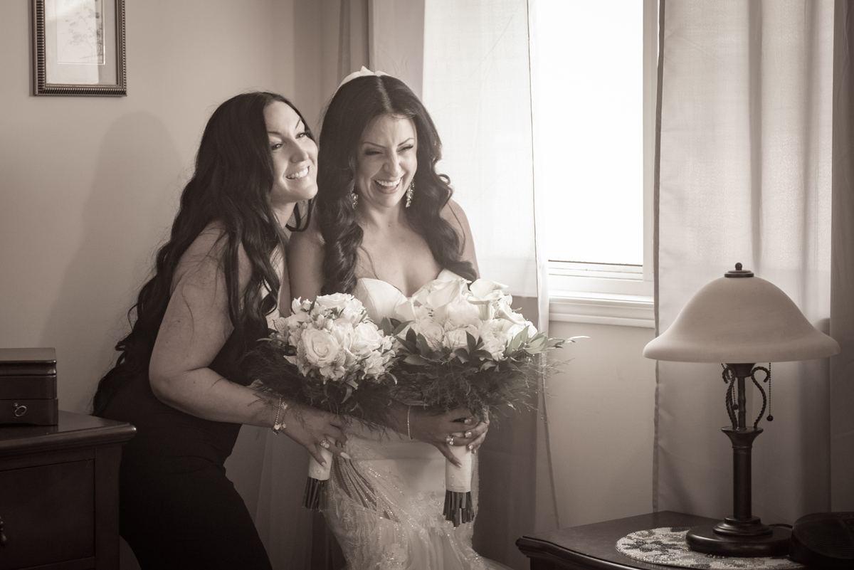 A bride in a white gown laughs joyfully with a smiling friend beside her, illuminated by soft window light.