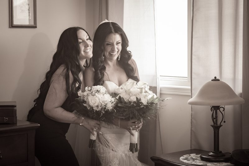 A bride in a white gown laughs joyfully with a smiling friend beside her, illuminated by soft window light.