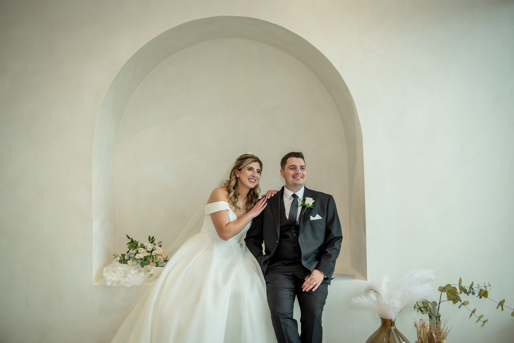 Bride and groom smile warmly together beneath an arched alcove, surrounded by delicate floral arrangements.