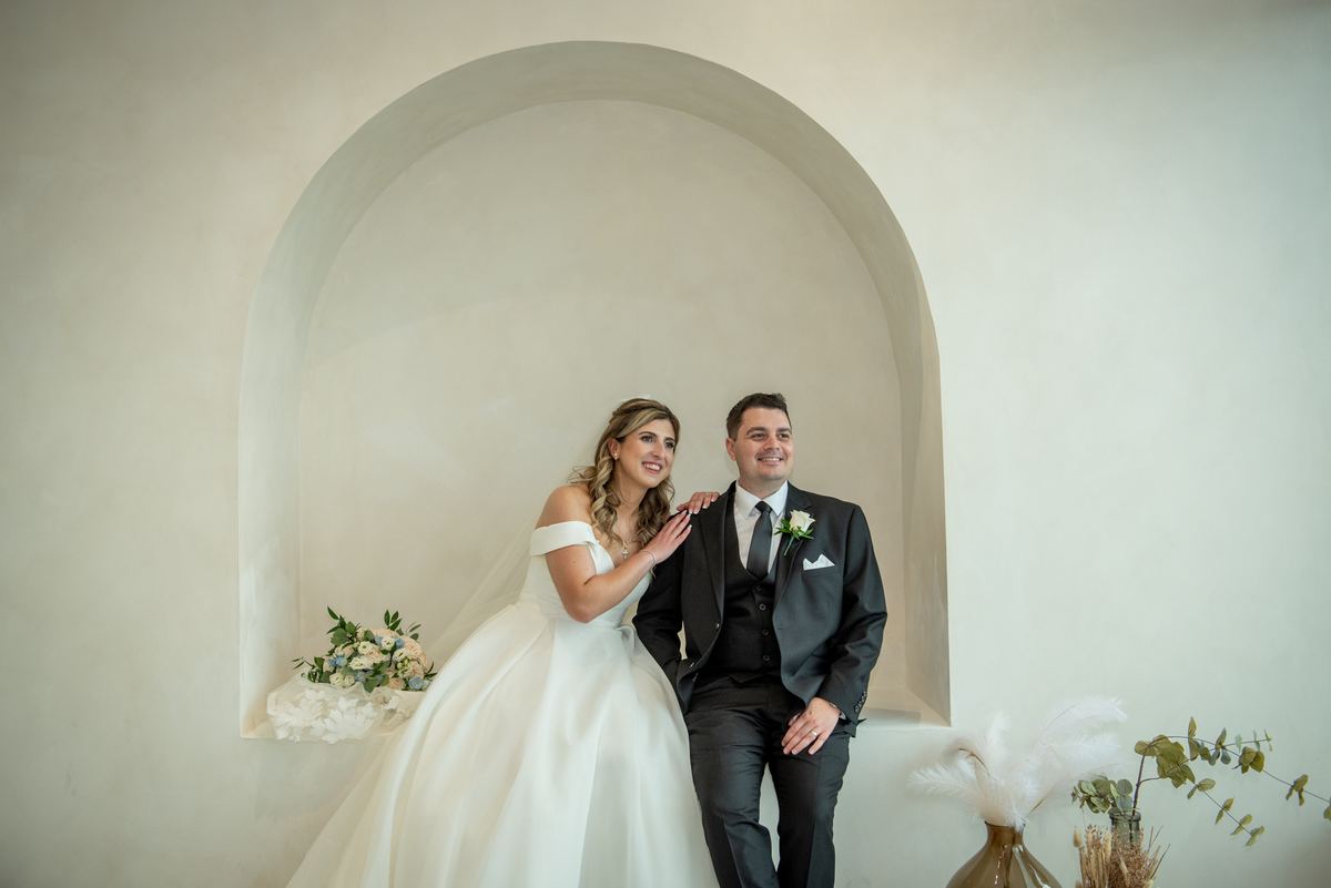 Bride and groom smile warmly together beneath an arched alcove, surrounded by delicate floral arrangements.