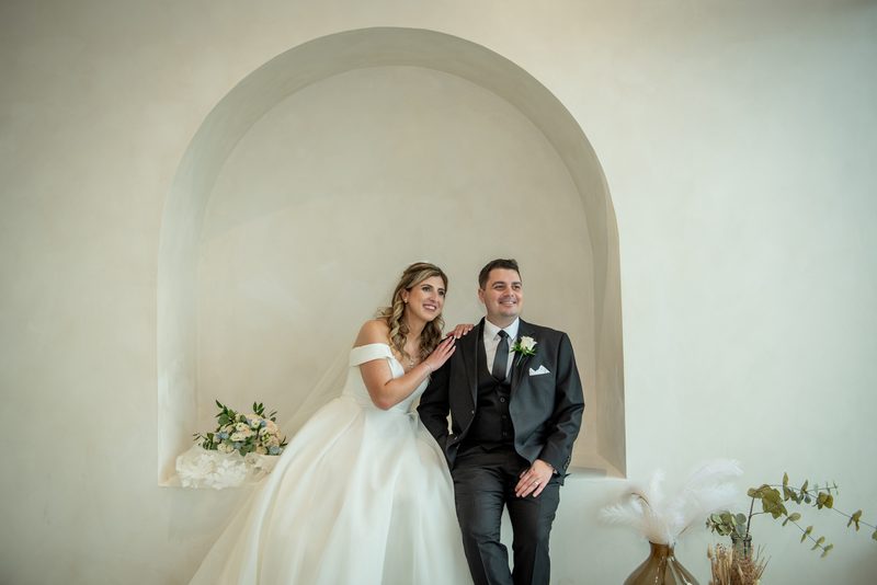 Bride and groom smile warmly together beneath an arched alcove, surrounded by delicate floral arrangements.