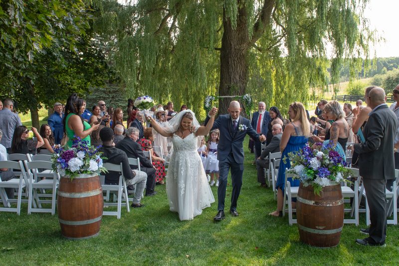 A smiling bride and groom hold hands as they walk down a grassy aisle under a weeping willow, surrounded by guests.