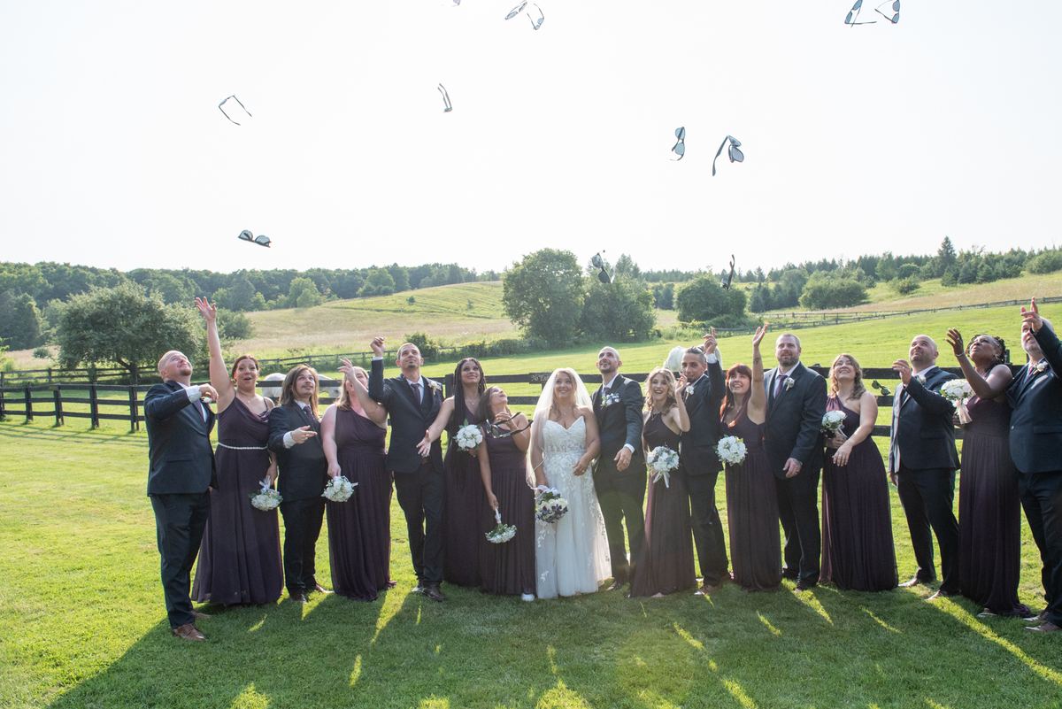 A wedding party stands outdoors, tossing bouquets into the air with joy as sunlight casts long shadows.