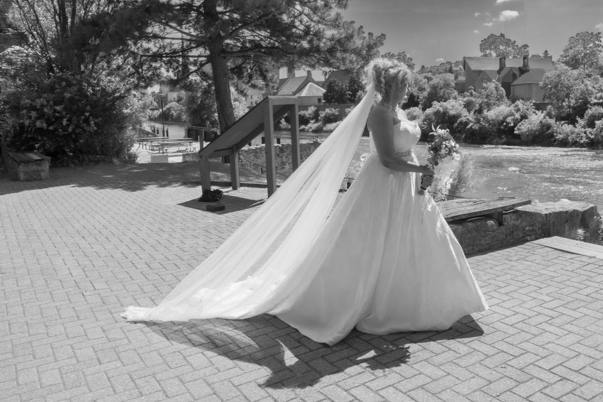 A bride in a white gown and veil stands by a river with trees and houses behind her, posing for a photo.
