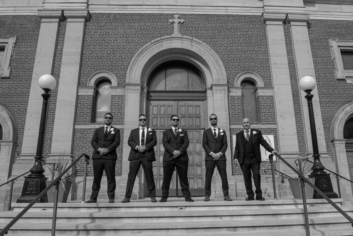 Five formally dressed men stand poised with clasped hands and sunglasses on stone steps outside a brick church.