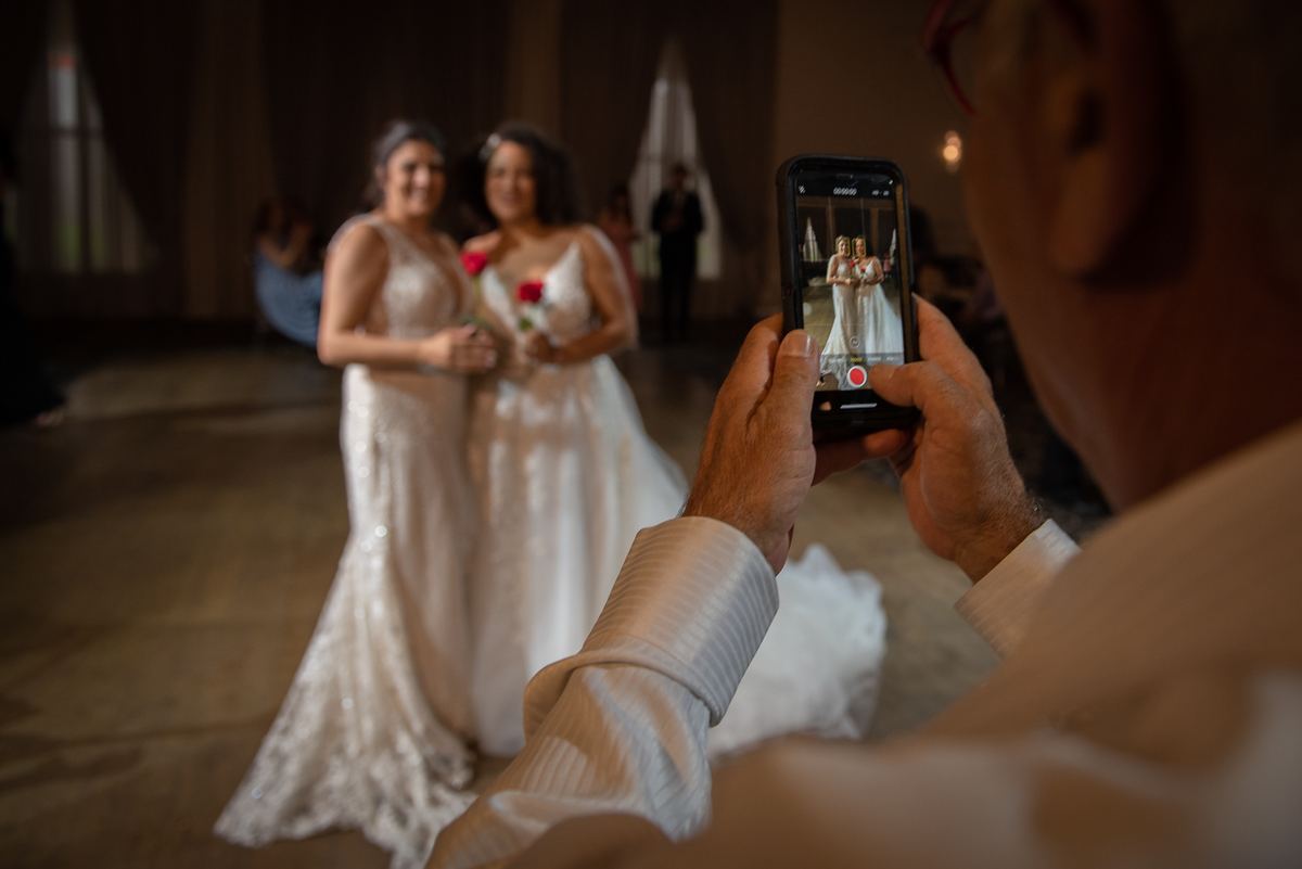 A guest films two brides dancing joyfully in a warmly lit ballroom, their gowns shimmering under the golden glow.