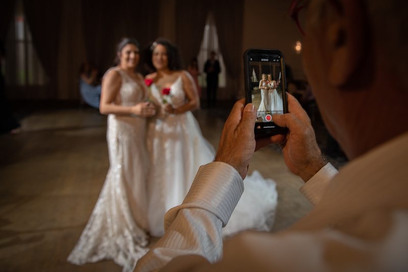 A guest films two brides dancing joyfully in a warmly lit ballroom, their gowns shimmering under the golden glow.