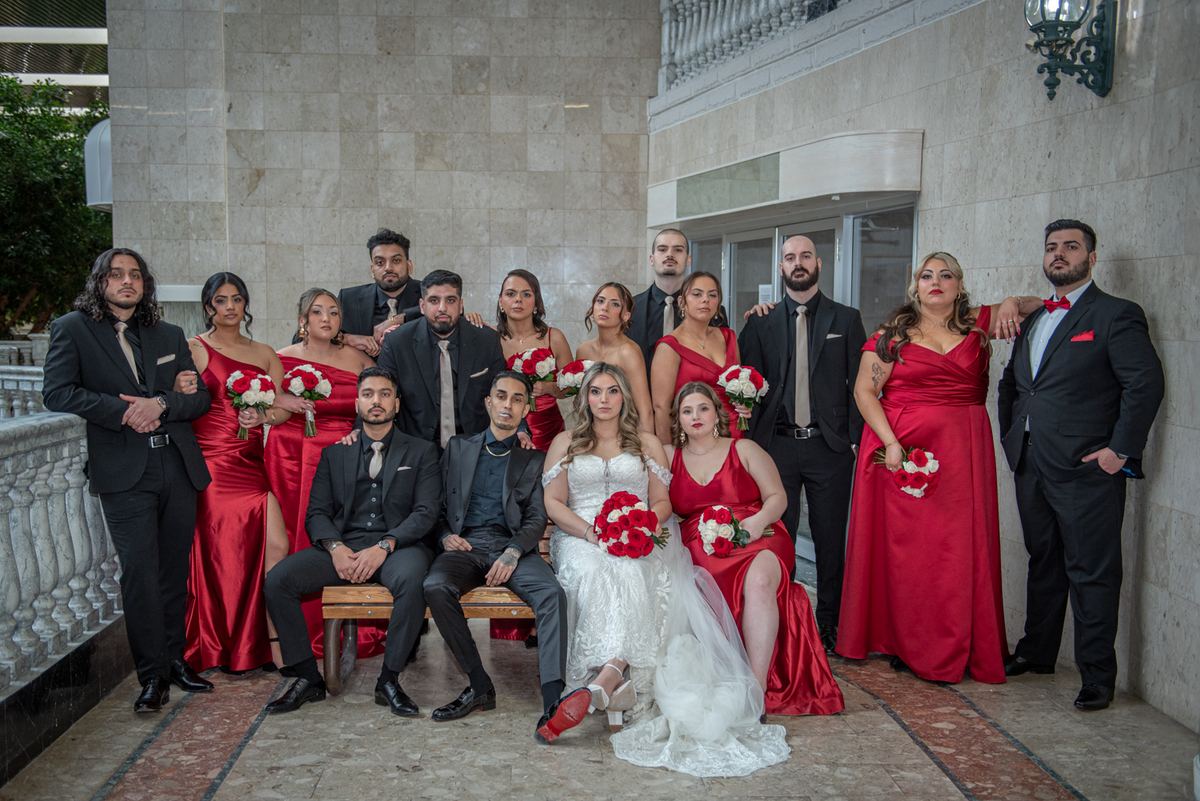 Bride in white gown sits center with groom and wedding party indoors, all holding red-and-white bouquets.