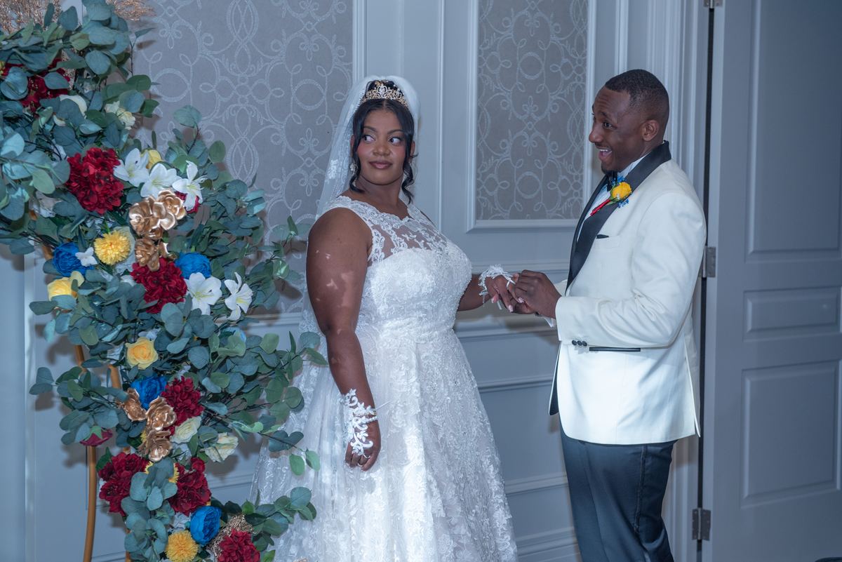 Bride and groom exchange vows, holding hands and gazing at each other affectionately under an ornate floral arch.