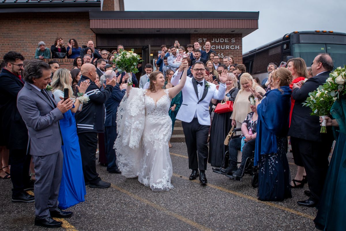 A smiling bride and groom exit a church, waving and holding hands as guests cheer with their phones.