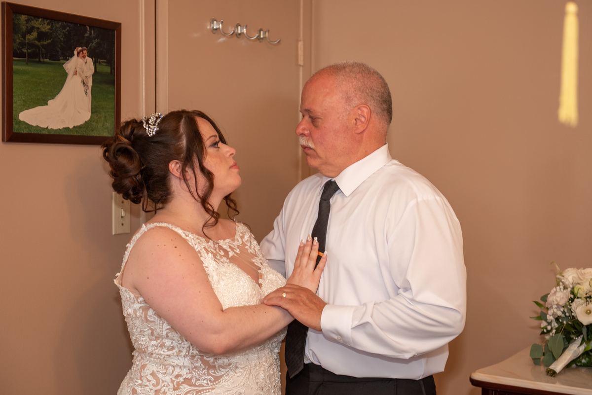 A bride in a lace gown looks up at her father, who holds her hand with love and pride beside a framed portrait of a bride.