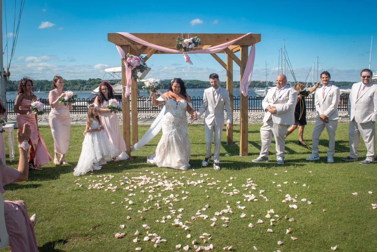 Bride and groom walk hand-in-hand under wooden arch adorned with pink drapery and flowers, surrounded by wedding party.