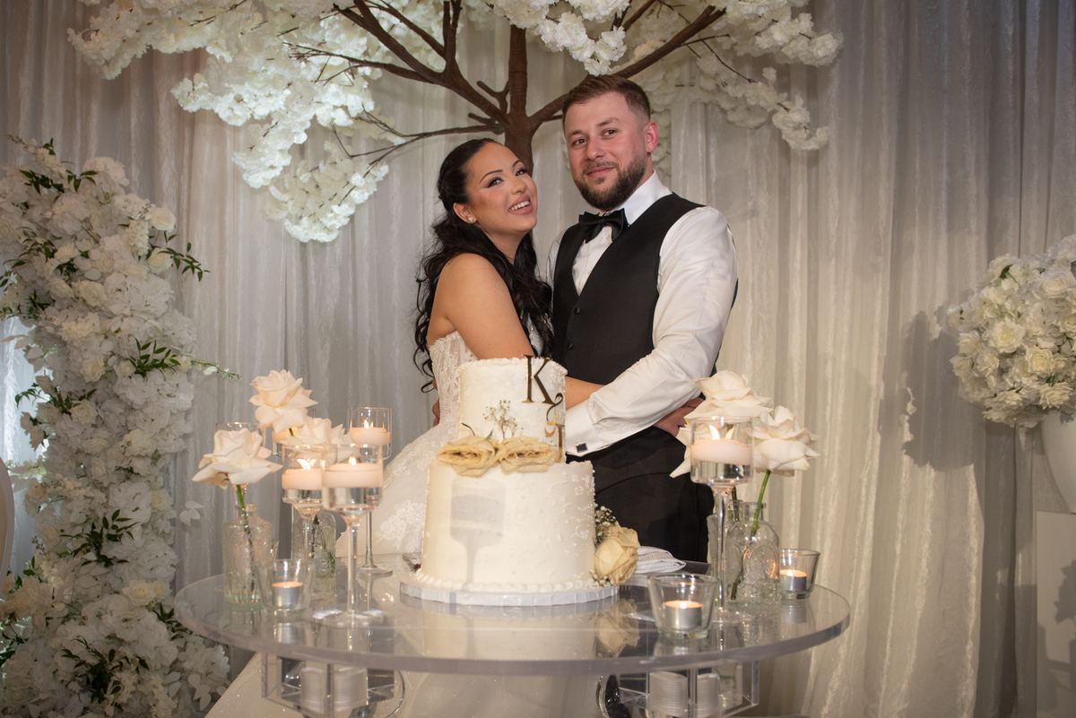 A smiling newlywed couple stands beside their wedding cake, decorated with candles and flowers, in front of white drapery.