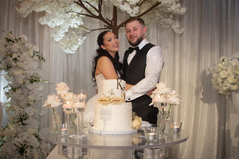 A smiling newlywed couple stands beside their wedding cake, decorated with candles and flowers, in front of white drapery.