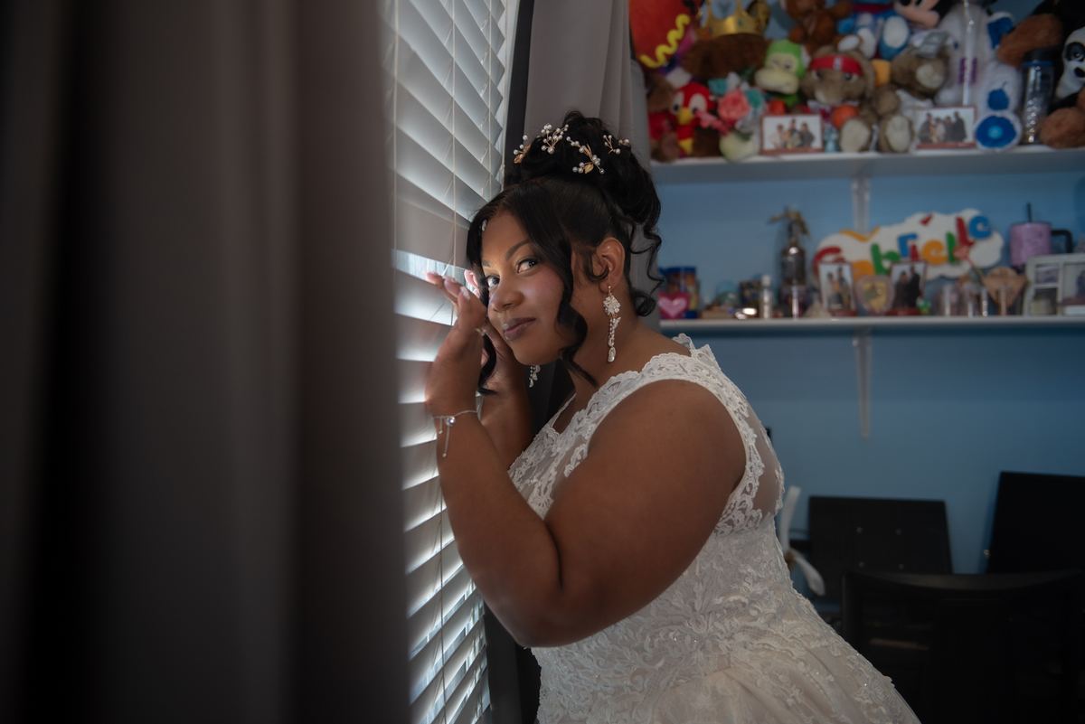 A bride in a white lace gown looks through window blinds, soft light highlighting her thoughtful expression.
