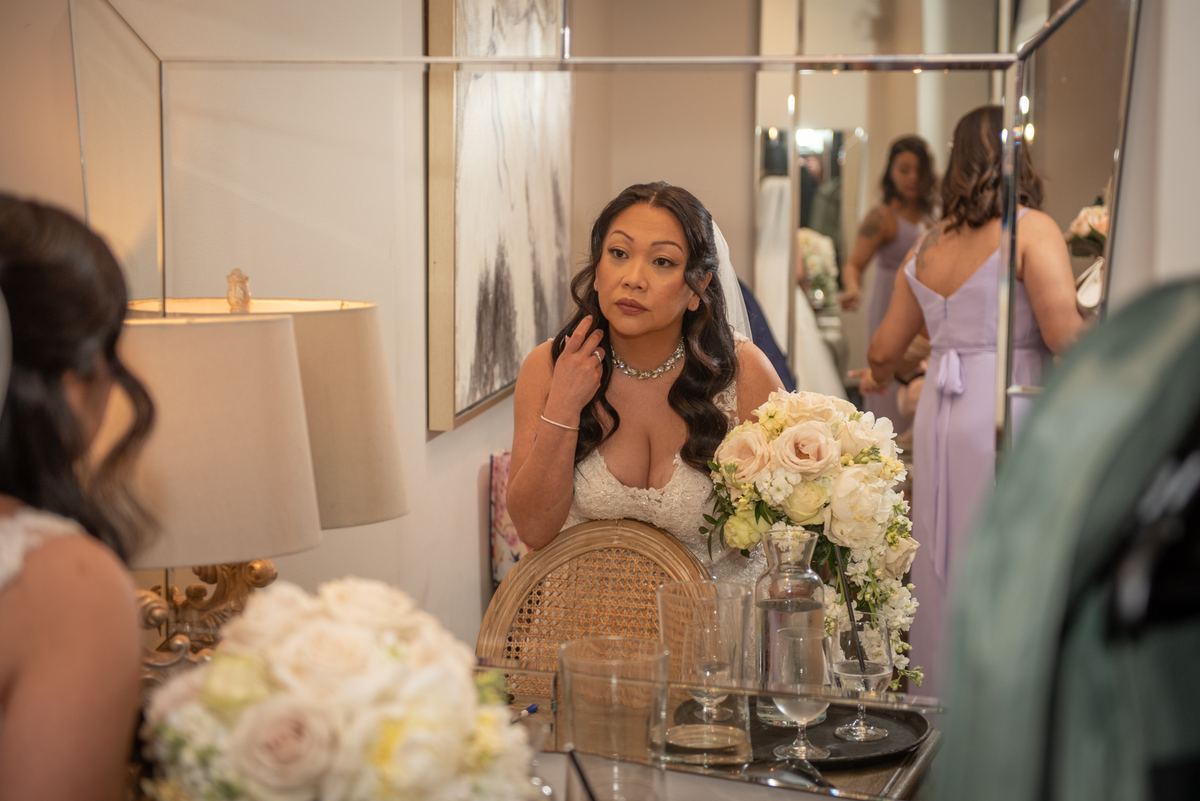 A bride in a white gown and veil adjusts her hair in front of a mirror, surrounded by floral arrangements.