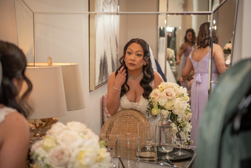 A bride in a white gown and veil adjusts her hair in front of a mirror, surrounded by floral arrangements.