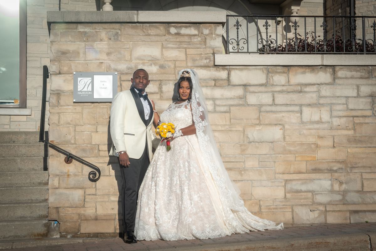 A bride in a lace gown and bouquet of yellow flowers stands with her groom in a white tuxedo near a stone wall.