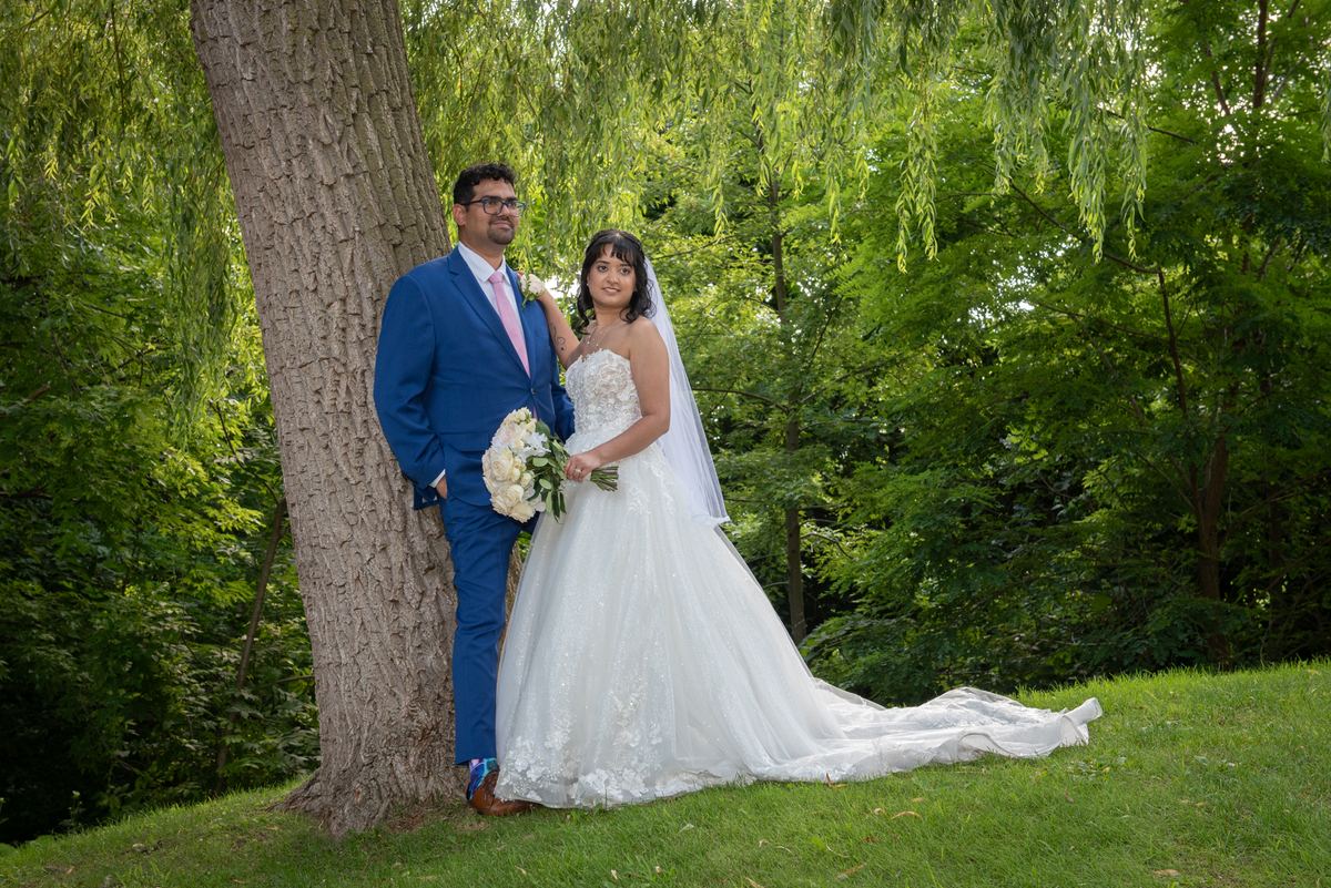 A newlywed couple poses under a willow tree in a park, the groom in a blue suit with a pink tie leaning against the tree.