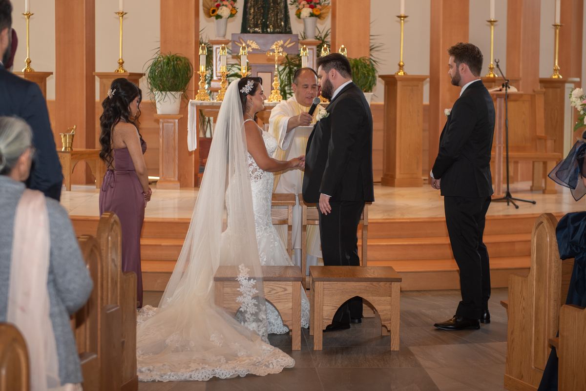 Bride and groom stand at the altar during a ceremony, illuminated by warm light with guests in pews and flowers on the altar.