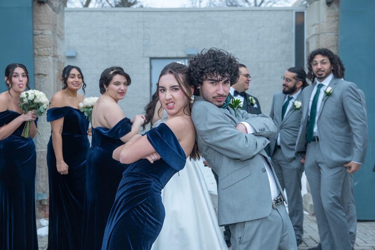 Bride and groom pose playfully with wedding party in the background, against a modern stone and blue wall backdrop.