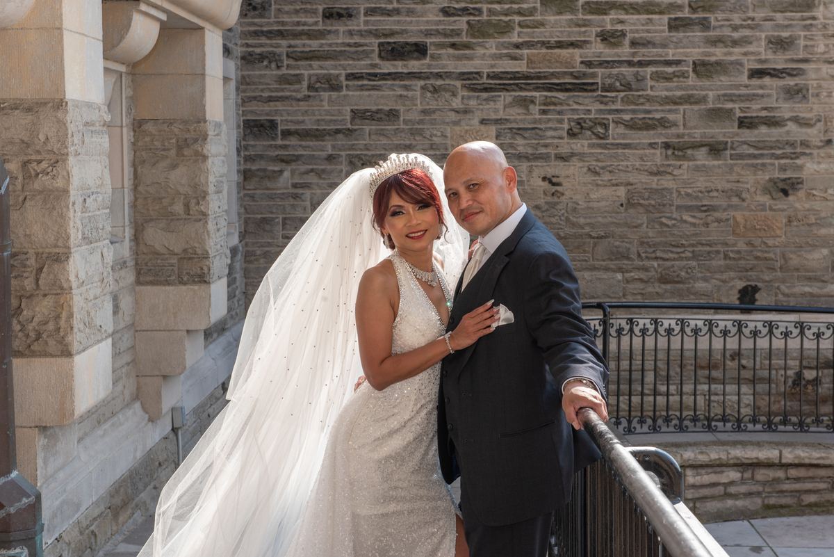 A newlywed couple in formal attire stands beside an ornate metal railing against a stone wall under bright daylight.