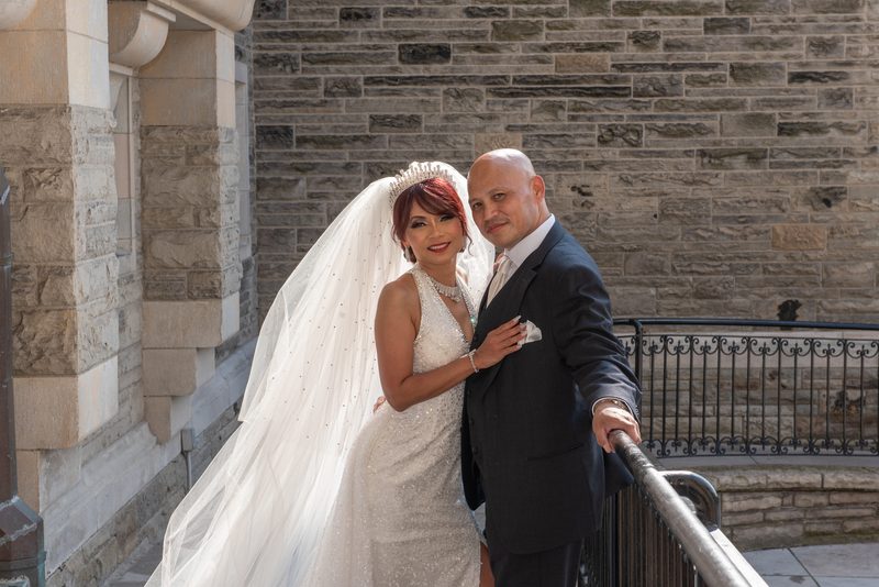 A newlywed couple in formal attire stands beside an ornate metal railing against a stone wall under bright daylight.