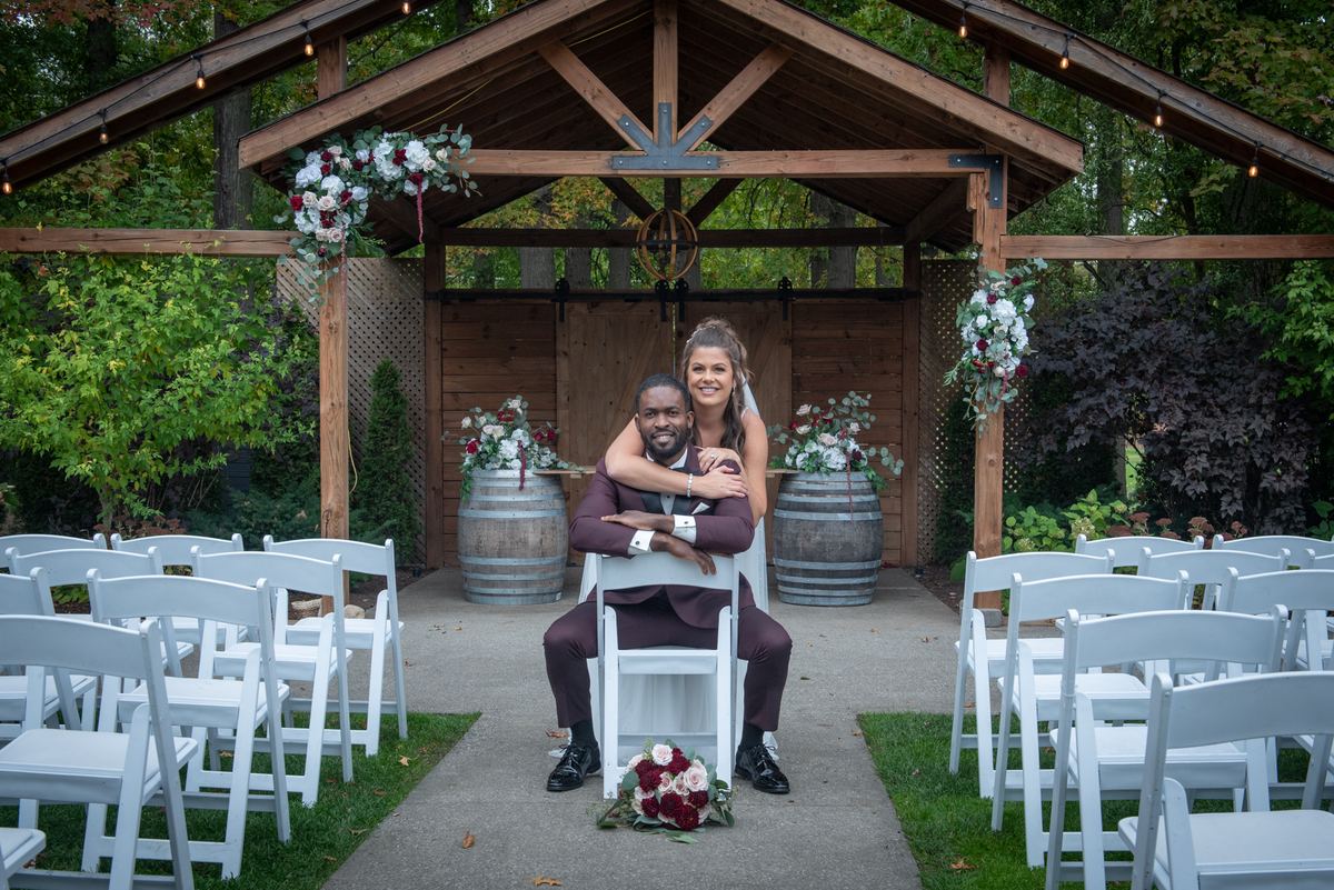 A smiling bride hugs her seated groom in an outdoor wedding aisle surrounded by white chairs and floral decorations.